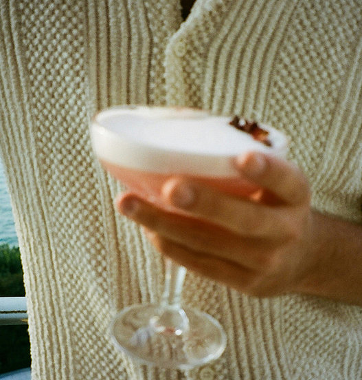 Person hält ein Cocktailglas mit einem hellen Drink und Schaumkrone vor einem beigen Strickoberteil.