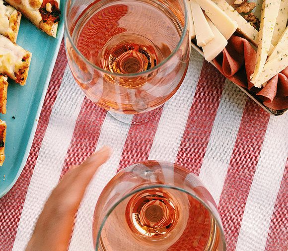 Hier schaut man von oben auf zwei Weingläser, gefüllt mit einem Roséwein. Die Gläser befinden sich auf einer rot und weiß gestreiften Decke. Links und oberhalb sind Speisen leicht angeschnitten. Man kann einen Käse erkennen. 