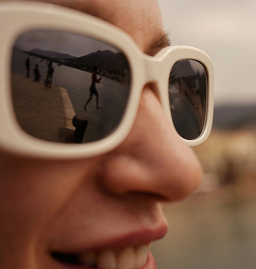 Detailaufnahme einer Person mit heller Sonnenbrille, in deren Gläsern sich eine Uferlandschaft spiegelt.