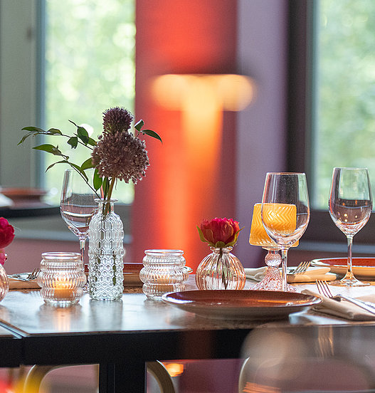 Stilvoll gedeckter Restauranttisch mit Gläsern, Blumenvasen und warmem Licht im Hintergrund.