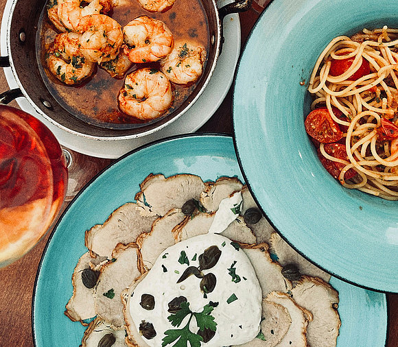 Auf dem Bild sind mehrere Speisen abgebildet. Man kann eine kleine Pfanne mit Garnelen erkennen, ein Pastagericht mit Tomaten sowie ein Vitello Tonnato. 