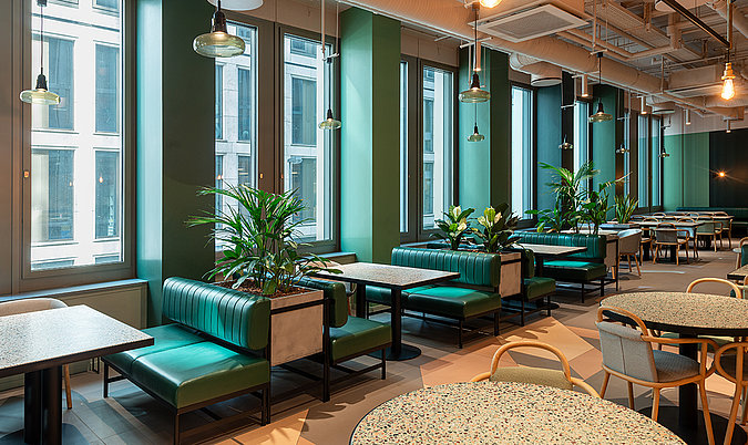 Fensterlounge mit Sitzbänken, Pflanzen und modernem Restaurantambiente entlang einer großzügigen Fensterfront.