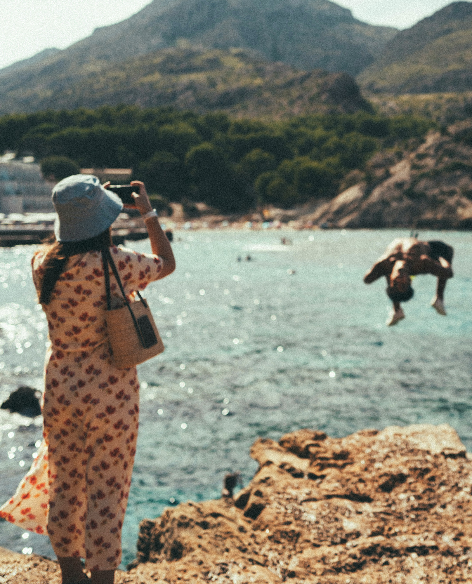 Person fotografiert eine Badebucht, während eine weitere Person von einem Felsen ins türkisfarbene Wasser springt.